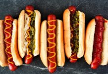 Dünya mutfağında geniş bir yer edinen hot dog, özellikle Amerika Birleşik Devletleri’nde adeta bir sembol haline gelmiştir. Kökeni 19. yüzyılın sonlarına kadar uzanan bu yiyecek, Almanya’dan Amerika’ya göç edenlerin beraberinde getirdiği sosis kültürü ile ortaya çıkmıştır. Uzun ekmek arasına yerleştirilen sosisin seneler içinde farklı soslarla buluşması, hot dog’u bugünkü popüler formuna kavuşturmuştur. Konulu bir haber görseli.