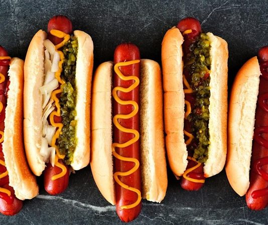 Dünya mutfağında geniş bir yer edinen hot dog, özellikle Amerika Birleşik Devletleri’nde adeta bir sembol haline gelmiştir. Kökeni 19. yüzyılın sonlarına kadar uzanan bu yiyecek, Almanya’dan Amerika’ya göç edenlerin beraberinde getirdiği sosis kültürü ile ortaya çıkmıştır. Uzun ekmek arasına yerleştirilen sosisin seneler içinde farklı soslarla buluşması, hot dog’u bugünkü popüler formuna kavuşturmuştur. Konulu bir haber görseli.