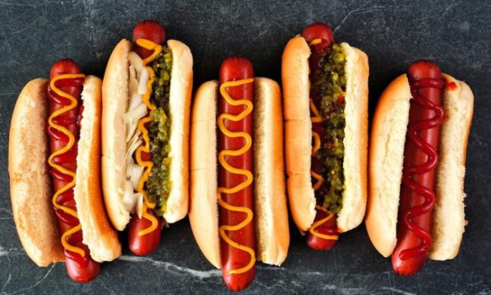 Dünya mutfağında geniş bir yer edinen hot dog, özellikle Amerika Birleşik Devletleri’nde adeta bir sembol haline gelmiştir. Kökeni 19. yüzyılın sonlarına kadar uzanan bu yiyecek, Almanya’dan Amerika’ya göç edenlerin beraberinde getirdiği sosis kültürü ile ortaya çıkmıştır. Uzun ekmek arasına yerleştirilen sosisin seneler içinde farklı soslarla buluşması, hot dog’u bugünkü popüler formuna kavuşturmuştur. Konulu bir haber görseli.